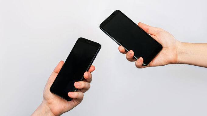 Two Hands Of Two Women Holding Mobile Phones With Blank Black Screens, Empty Copy Space For Design Isolated On A Gray Background. Internet Connection, File Transfer Via Bluetooth, Information Exchange