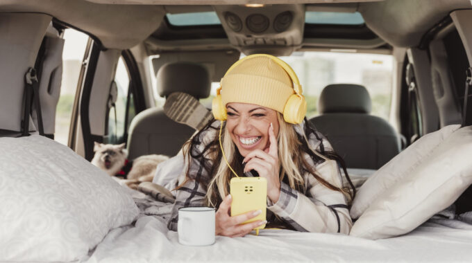 A Laughing Woman Laying Indoor Mini Camper Van Using A Mobile Phone And Listening To The Music With Yellow Headphones. Travel Vanlife Digital Nomad People