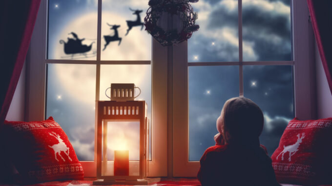 Merry Christmas And Happy Holidays! Cute Little Child Girl Sitting By Window And Looking At Santa Claus Flying In His Sleigh Against Moon Sky. Room Decorated On Christmas. Kid Enjoy The Holiday.
