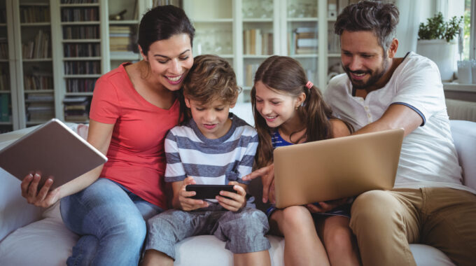Happy Family Sitting On Sofa And Using Laptop, Mobile Phone And Digital Tablet At Home