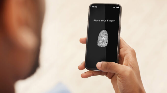 Biometric Identification Concept. Over The Shoulder View Of Black Man Holding Mobile Phone In Hand, Showing App For Fingerprint Scanning With A Zone To Touch With Thumbprint Icon On The Device Screen